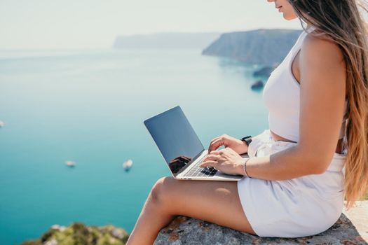 Successful business woman in yellow hat working on laptop by the sea. Pretty lady typing on computer at summer day outdoors. Freelance, travel and holidays concept.