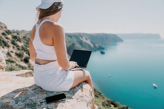 Successful business woman in yellow hat working on laptop by the sea. Pretty lady typing on computer at summer day outdoors. Freelance, travel and holidays concept.