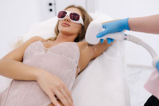 Beautician doing epilation on female's armpit in beauty center. Woman receiving laser light hair removal treatment for hairless smooth skin at cosmetology salon.