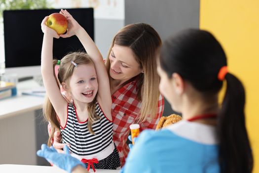 A little girl with her mother at the doctor rejoices at an apple, close-up. Consultation with a pediatric nutritionist, natural and synthetic vitamins