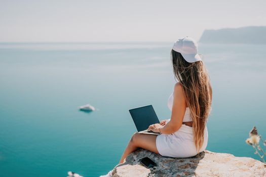 Successful business woman in yellow hat working on laptop by the sea. Pretty lady typing on computer at summer day outdoors. Freelance, travel and holidays concept.