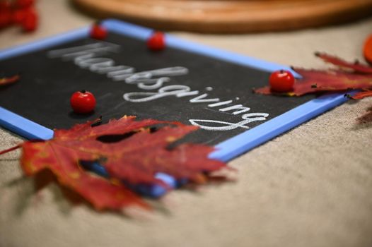 Selective focus on chalk lettering Thanksgiving Day on the black board with fallen autumn maple leaves and viburnum berries, lying down on a linen tablecloth. Autumnal still life