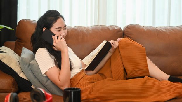 Happy young woman in casual clothes resting on sofa ai bright living room and talking on mobile phone with her friends.