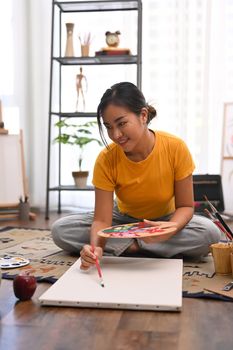 Millennial beautiful woman painting picture on canvas with oil paints in bright home studio. Leisure activity and art concept.