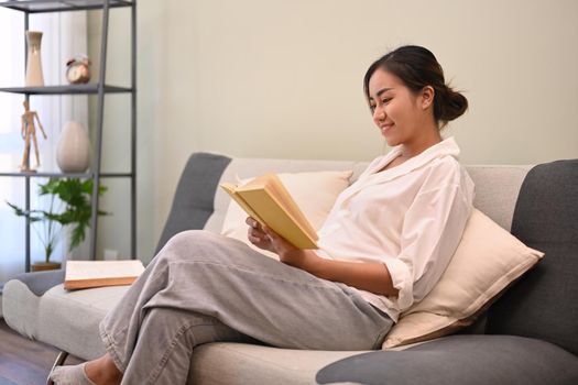 Peaceful asian woman resting on sofa and reading book, spending leisure time at home.