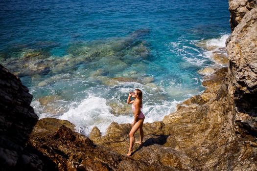 Young beautiful woman in a swimsuit stands on a rocky beach of the Mediterranean Sea. The concept of sea recreation. Selective focus