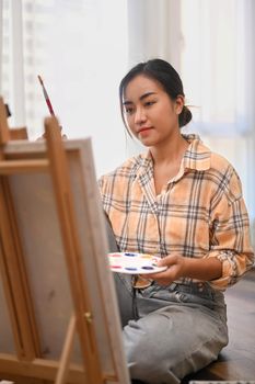 Positive young female artist holding palette and painting picture on canvas with oil paints in her home studio.