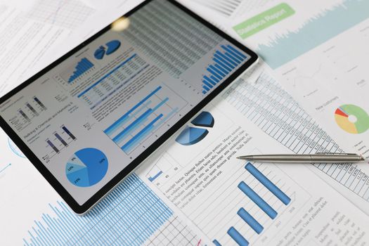 On statistical documents lies a tablet with charts on the screen, close-up. Benchmarking, financial performance, audit
