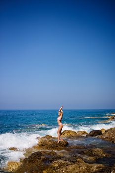 Beautiful young woman in a swimsuit on a rocky beach on a sunny day against the backdrop of waves. Vacation in the summer season. Selective focus