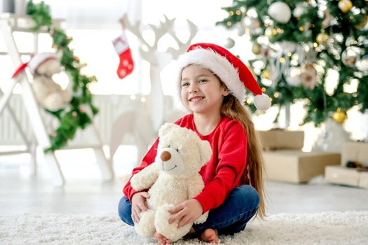 Happy child girl hugging teddy bear toy in Christmas time in room with decorated tree and gifts. Pretty kid with presents at home in New Year celebration
