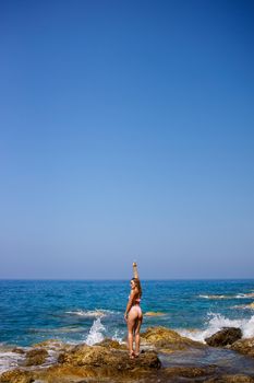 Beautiful young woman in a swimsuit on a rocky beach on a sunny day against the backdrop of waves. Vacation in the summer season. Selective focus