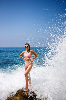 Beautiful young woman in a swimsuit on a rocky beach on a sunny day against the backdrop of waves. Vacation in the summer season. Selective focus