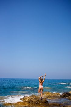 Beautiful young woman in a swimsuit on a rocky beach on a sunny day against the backdrop of waves. Vacation in the summer season. Selective focus