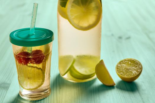 bottle and glass with lid and cane with cold water and slices of lime, lemon and red berries, illuminated by sunlight on a green wooden table with some pieces of citrus, summer refreshments background