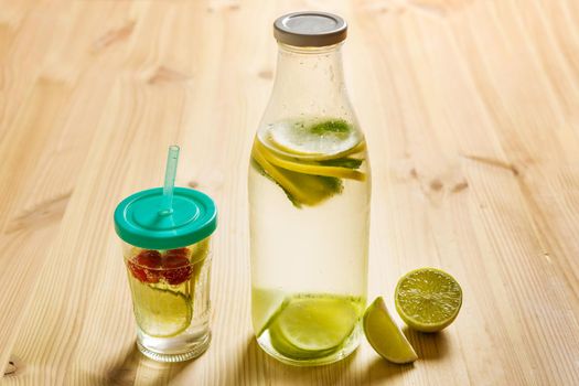 glass bottle and glass with lid and cane, with cold water and slices of lemon, lime, berries and mint, are illuminated by sunlight on a wooden table with some pieces of citrus