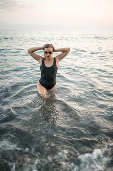 Attractive young woman in sunglasses comes out from the sea at sunset light in a black swimsuit. Selective focus