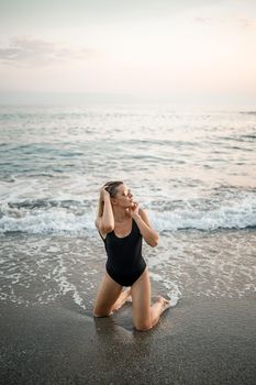 Attractive young woman in sunglasses kneels in the sea at sunset light in a black swimsuit. Selective focus