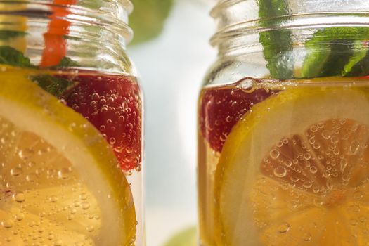 close-up of two glass jars illuminated by sunlight with refreshing cold lemonade water, lemon slices, red berries, mint leaves and drinking cane. Summer citrus soda background