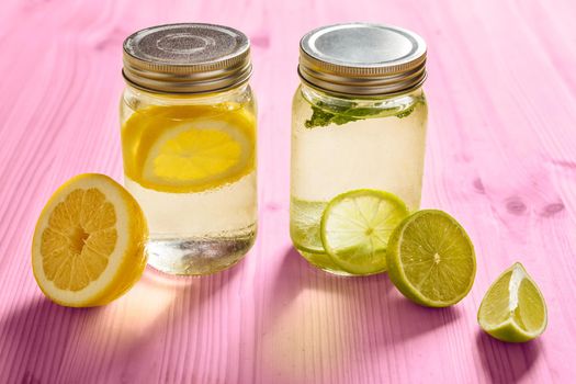 two glass jars with lid, one with cold water and slices of lemon and another with slices of lime and mint, are illuminated by sunlight and on a pink wooden table with some pieces of citrus fruits