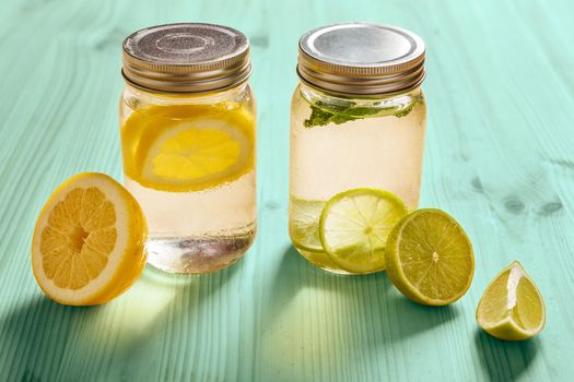 two glass jars with lid, one with cold water and slices of lemon and another with slices of lime and mint, are illuminated by sunlight and on a turquoise wooden table with some pieces of citrus fruits