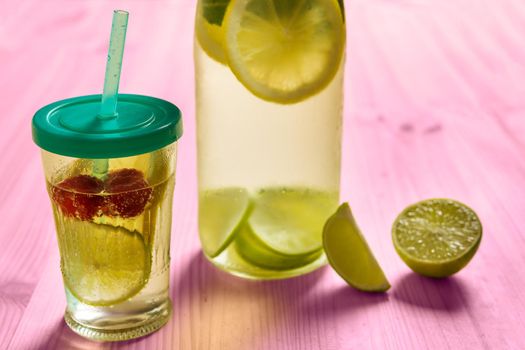 bottle and glass with lid and cane with cold water and slices of lime, lemon and red berries, illuminated by sunlight on a pink wooden table with some pieces of citrus, summer refreshments background