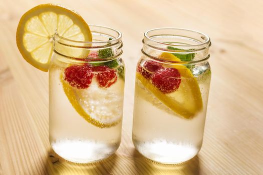 two glass jars with cold water, slices of lemon, mint and red berries, illuminated by sunlight on a wooden table with some pieces of citrus, summer refreshments background, copy space