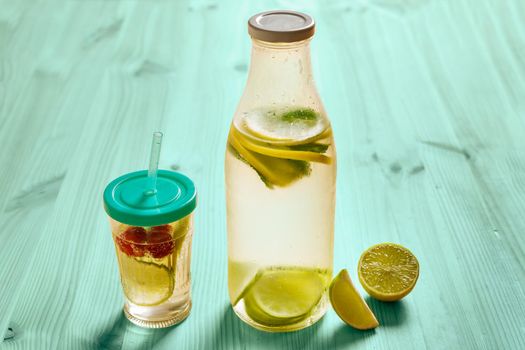 glass bottle and glass with lid and cane, with cold water and slices of lemon, lime, berries and mint, are illuminated by sunlight on a turquoise wooden table with some pieces of citrus