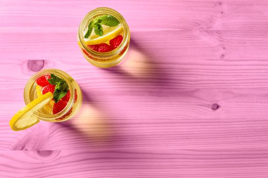 overhead shot of two glass jars with cold water, lemon slices, red berries and mint on one side of a pink wooden table illuminated by sunlight. Summer refreshment background with citrus, copy space