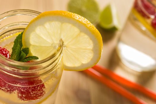 glass jar lit by sunlight with refreshing cold lemonade water, lemon slices, red berries and mint leaves on a unfocused wooden table with drink cane and pieces of citrus. Summer refreshment background