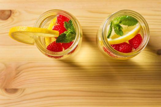 overhead shot of two glass jars with cold water, lemon slices, red berries and mint leaves on a wooden table, are illuminated by sunlight. Summer refreshment background with citrus, copy space