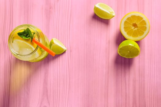 glass jar with homemade lemonade, mint and a cane, on the pink wooden table there are also wedges of lime and lemon