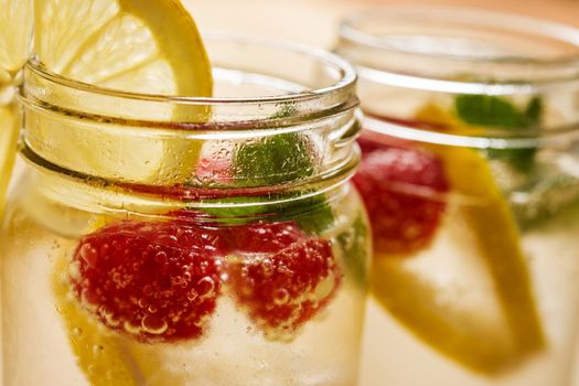 close-up of a glass jar with cold water, slices of lemon, mint and red berries, illuminated by sunlight on a wooden table, in the background it looks out of focus another glass equal