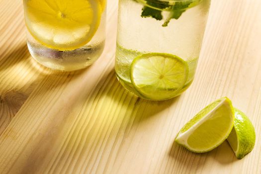 cold summer refreshments with slices of citrus and mint leaves, are on a wooden table and illuminated by sunlight, there are also lemon wedges and copy space