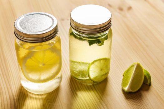 two glass jars with lids filled with water with slices of lime and lemon, are on a wooden table and illuminated by sunlight, there are also two slices of lime on the table