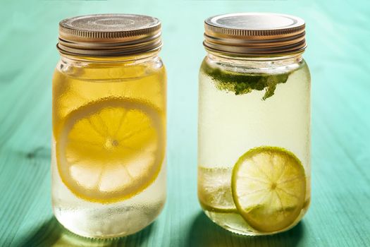 two glass jars with lids on a turquoise wooden table illuminated by sunlight, one has cold water with lemon and the other with lime and mint