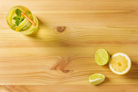 flat lay of homemade lemonade with mint and a cane to drink in a glass jar lit by sunlight, it's on a wooden table, there are also a few pieces of lime and lemon and copy space