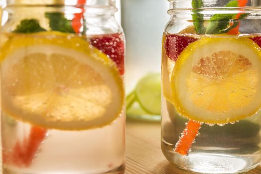 close-up of two glass jars illuminated by sunlight with refreshing cold lemonade water, lemon slices, red berries, mint leaves and drink canes. Summer citrus soda background