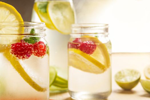 Two glass jars and a bottle with refreshing cold water, lemon slices, red berries and mint on a table with pieces of citrus lit by sunlight. Summer refreshment background. copy space