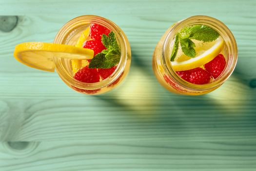 overhead shot of two glass jars with cold water, lemon slices, red berries and mint leaves on a green wooden table, are illuminated by sunlight. Summer refreshment background with citrus, copy space