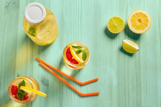 top view, two glass jars and a bottle with refreshing cold lemonade water, lemon, red berries and mint on a green wooden table with citrus lit by sunlight, Summer refreshment background Copy space