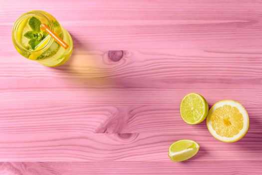 flat lay of homemade lemonade with mint and a cane to drink in a glass jar lit by sunlight, it's on a pink wooden table, there are also a few pieces of lime and lemon and copy space