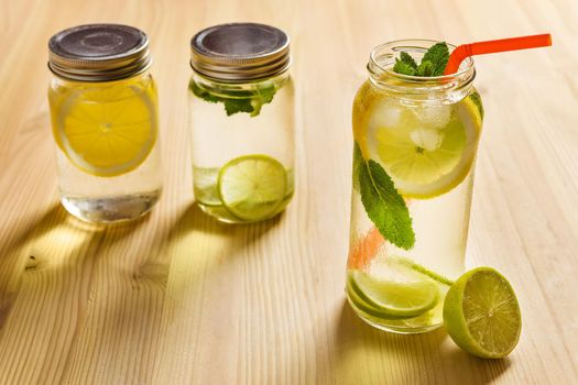 Summer refreshments with slices of lime, lemon and mint leaves illuminated by sunlight. There are two glass jars with lids and one with a cane to drink, these are on a wooden table