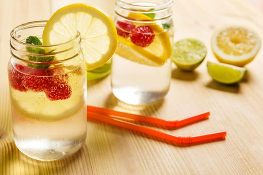 Two glass jars with refreshing cold water, lemon slices, red berries and mint on a wooden table with drinking canes and slices of citrus lit by sunlight. Summer refreshment background. copy space