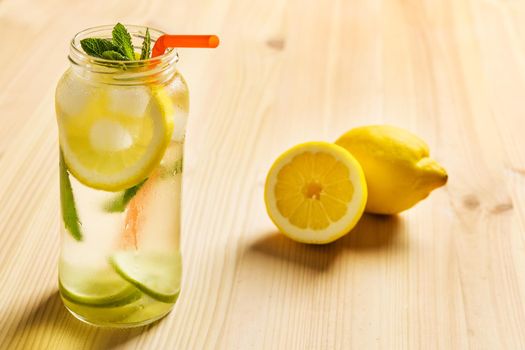 lemons on a wooden table next to a lemonade in a glass jar, the jar contains water, slices of lemon and lime, ice, mint leaves and a cane to drink