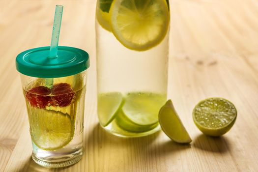 bottle and glass with lid and cane with cold water and slices of lime, lemon and red berries, illuminated by sunlight on a wooden table with some pieces of citrus, summer refreshments background