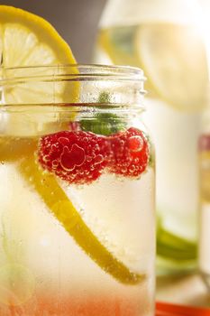 vertical photo of a glass jar with cold water, slices of lemon, red berries and mint with a bottle with citrus pieces illuminated by sunlight in the background. Summer refreshment with citrus