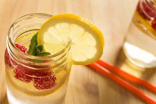 glass jar illuminated by sunlight with refreshing cold lemonade water, lemon slices, red berries and mint leaves on a wooden table with drink canes. Summer citrus soda background. copy space