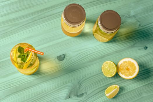 Flat Lay of lemonade with mint and a cane to drink in a glass jar illuminated by sunlight, is on a turquoise wooden table with two other jars with lid, some pieces of lemon and lime and copy space