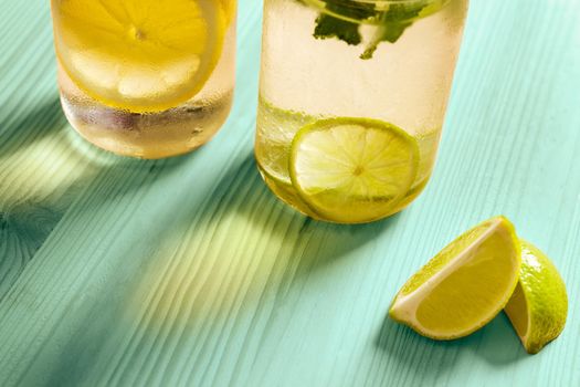 cold summer refreshments with slices of citrus and mint leaves, are on a turquoise wooden table and illuminated by sunlight, there are also lemon wedges and copy space