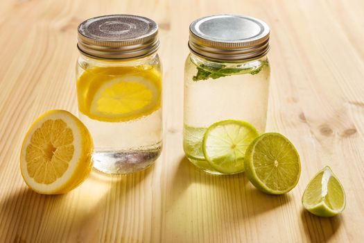 two glass jars with lid, one with cold water and slices of lemon and another with slices of lime and mint, are illuminated by sunlight and on a wooden table with some pieces of citrus fruits